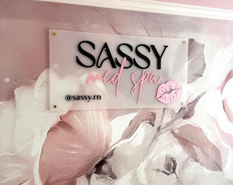 May include: A clear acrylic sign displaying the words "SASSY med spa" in black and pink, with a pink lip graphic. The sign is mounted on a floral wallpaper and includes the Instagram handle @sassy.rn.