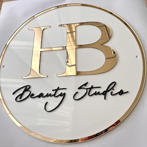 May include: A white circular sign with a gold border and gold letters "HB" in the center. The text "Beauty Studio" is written in black cursive below the letters.