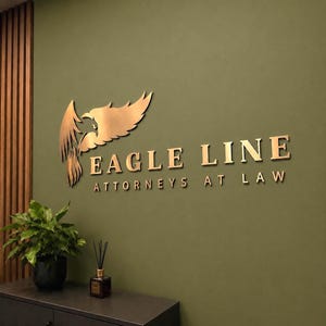 May include: A three-dimensional wall sign featuring a metallic gold eagle emblem and the text EAGLE LINE ATTORNEYS AT LAW in a serif font. The sign is mounted on a solid olive green wall, positioned above a dark surface with a plant and diffuser.