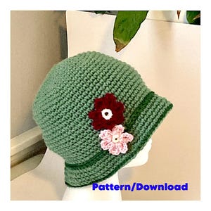 Könnte beinhalten: Ein grüner Häkel-Cloche-Hut mit breiter Krempe und einer dekorativen Blumenverzierung. Die Blume ist aus Häkelgarn in den Farben Rosa, Rot und Weiß gefertigt.