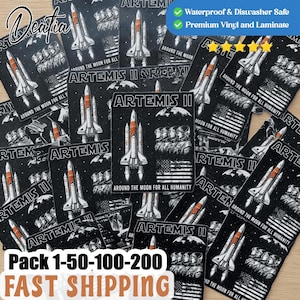 Puede incluir: Una colección de pegatinas Artemis II con un cohete, la bandera estadounidense y el texto "AROUND THE MOON FOR ALL HUMANITY". Las pegatinas son negras con detalles en blanco y naranja. La imagen también incluye el texto "Pack 1-50-100-200".