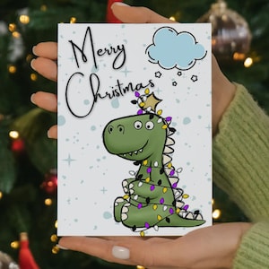 Może przedstawiać: Kartka świąteczna z kreskówkowym zielonym dinozaurem ozdobionym lampkami choinkowymi. Na kartce widnieje napis "Merry Christmas" w fantazyjnej czcionce, z jasnoniebieską chmurką i gwiazdkami nad dinozaurem.