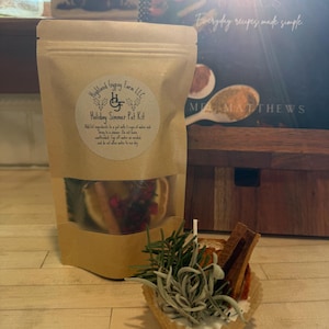 May include: A kraft paper bag labeled "Highland Gypsy Farm LLC Holiday Simmer Pot Kit" with a clear window showing ingredients. A small cupcake liner holds rosemary, sage, cinnamon sticks, and other ingredients. A cookbook is in the background.