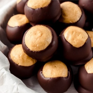 May include: A close-up shot of a pile of homemade Buckeye candies. Each candy is a ball of peanut butter fudge partially dipped in dark chocolate, with a light brown circle of peanut butter visible on top. The candies are arranged in a white paper-lined container.