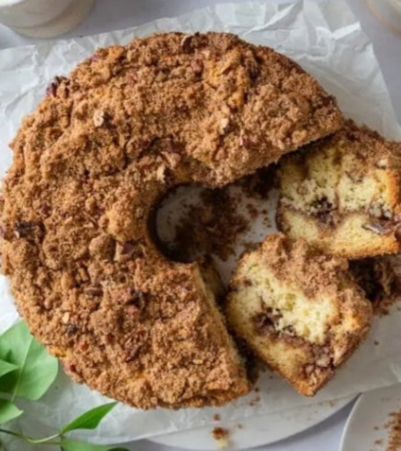 Puede incluir: Un primer plano de un pastel de caf&eacute; con cobertura de migas. El pastel tiene forma de anillo y se ha cortado en rebanadas. El pastel est&aacute; sobre una superficie blanca con papel de pergamino.
