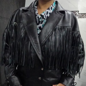 Puede incluir: Chaqueta de cuero negro con flecos en el pecho y las mangas. La chaqueta tiene un cuello con muescas, dos botones plateados y detalles de cordones en las mangas. La chaqueta se lleva sobre una camisa estampada.