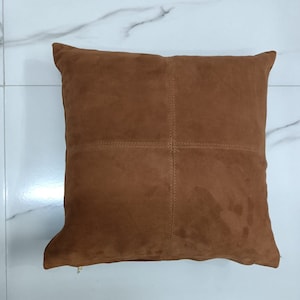 May include: A brown suede cushion with a zipper closure. The cushion is divided into four sections by stitching.