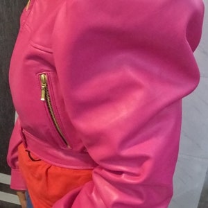 May include: A pink leather jacket with a cropped style and puffed sleeves. The jacket has a zipper closure and gold hardware.