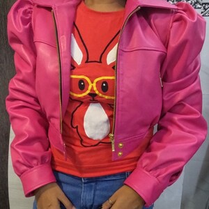 May include: A pink cropped leather jacket with puffed sleeves and a gold zipper. The jacket has a gold button detail on the front. The model is wearing a red t-shirt with a white bunny graphic and blue jeans.