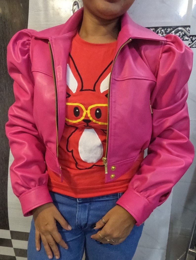 May include: A pink cropped leather jacket with puffed sleeves and a gold zipper. The jacket has a gold button detail on the front. The model is wearing a red t-shirt with a white bunny graphic and blue jeans.