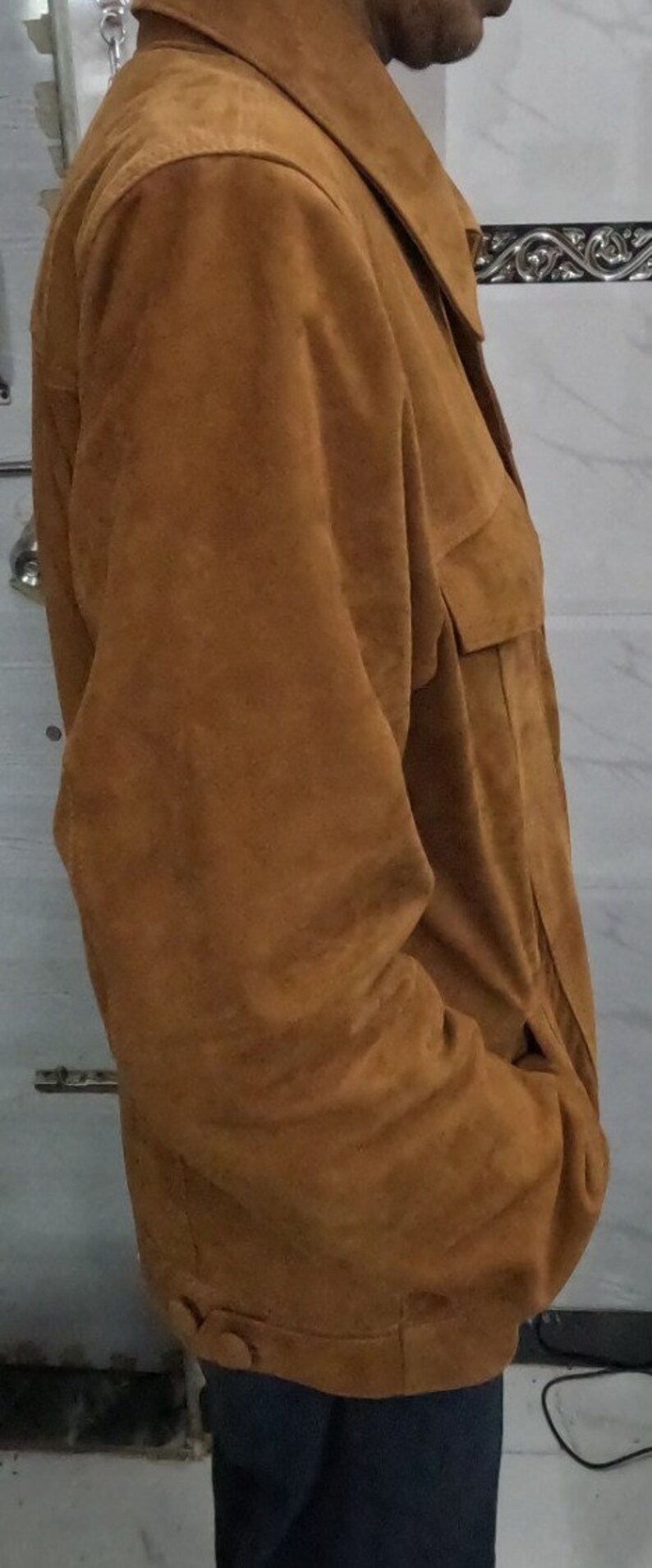 May include: A brown suede jacket with a collared neckline and a button closure. The jacket has a flap pocket on the left side and a button on the bottom right side.