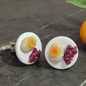 May include: A pair of cufflinks with a white ceramic base and a small, realistic depiction of a fried egg with a yolk and a side of purple jam.