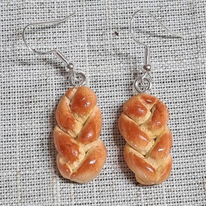 Puede incluir: Un par de pendientes de aro de plata con un diseño de pan trenzado. El pan es de color marrón claro y tiene un acabado brillante.