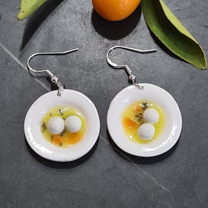 Peut inclure: Une paire de boucles d'oreilles en céramique blanche en forme de petites assiettes avec un bouillon jaune et des boulettes blanches. Les boucles d'oreilles sont attachées à des crochets argentés.
