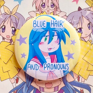 May include: A yellow button with a cartoon character with blue hair and a pink and white sailor uniform. The text on the button reads "BLUE HAIR AND PRONOUNS" with stars in the background.