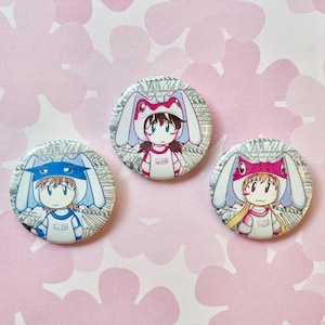 May include: Three round white button pins with cartoon characters wearing bunny ears. The characters are wearing different colored clothing and have different colored hair. The buttons are printed with a newspaper-like pattern.