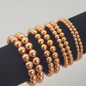 May include: A set of five copper beaded bracelets, stacked on a black display stand. The bracelets are made of shiny, round copper beads.