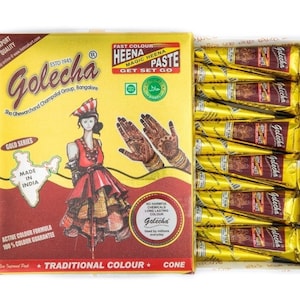 May include: A box of Golecha henna paste cones. The box is yellow and red with text and an illustration of a woman in traditional Indian attire. The box contains multiple gold-colored cones of henna paste, ready for use.