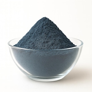 Op de afbeelding: Een heldere glazen kom gevuld met een berg donkerblauw poeder. Het poeder heeft een fijne, korrelige textuur en is hoog opgestapeld in het midden van de kom. De kom staat op een witte ondergrond.
