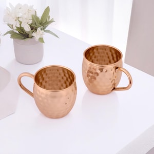May include: Two copper-colored Moscow Mule mugs with a hammered texture and curved handles. The mugs are sitting on a white surface, with a small vase of white flowers in the background. The mugs have a shiny, metallic finish.