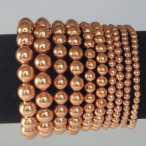 May include: A set of 10 copper bracelets, each with a different size of copper beads. The bracelets are stacked on top of each other, with the largest bracelet on the bottom and the smallest bracelet on top.