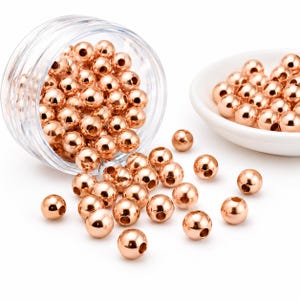 May include: Rose gold metal beads, spherical in shape, are scattered across a white surface. Some beads are contained in a clear plastic jar, while others are in a white ceramic dish. The beads have a polished, reflective surface.