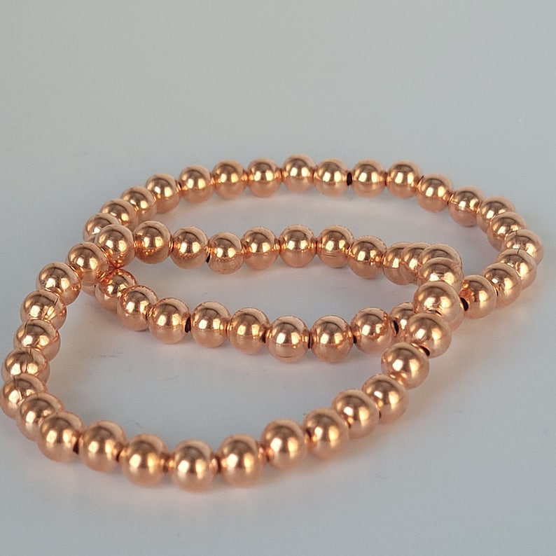 May include: Two copper-colored beaded bracelets, one stacked on top of the other. The bracelets are made of small, round beads.
