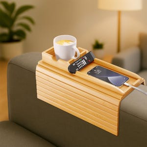 Puede incluir: Una bandeja de brazo de sofá de bambú con un portavasos incorporado, que sostiene una taza blanca con una bebida. Un mando a distancia negro y un teléfono inteligente también están en la bandeja. La bandeja está diseñada para el brazo del sofá.