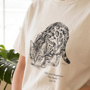 May include: Cream-colored t-shirt featuring a detailed black and white illustration of a Bengal cat in a stalking pose. The shirt includes text below the image: "Felis catus bengalensis - The Hunter - Sept 14."