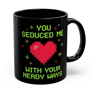 May include: Black ceramic mug with the text "YOU SEDUCED ME WITH YOUR NERDY WAYS" in green pixelated font. A red pixelated heart is in the center, with green and red pixelated accents. The mug has a black handle.