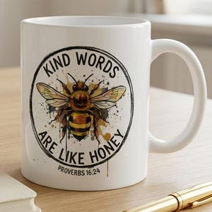 以下が含まれることがあります： 白いセラミックマグカップ。 蜂のイラストと「Kind Words Are Like Honey」と「Proverbs 16:24」の文字が描かれています。 マグカップは、ペン、開いた本、黄色い花の入った花瓶が置かれた木製のテーブルの上にあります。
