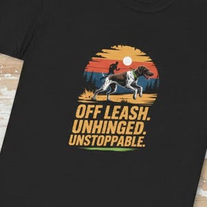 May include: Black t-shirt featuring a graphic of a dog running in front of a sunset. The text "OFF LEASH. UNHINGED. UNSTOPPABLE." is printed below the graphic in yellow and white. The shirt is made of a soft material.