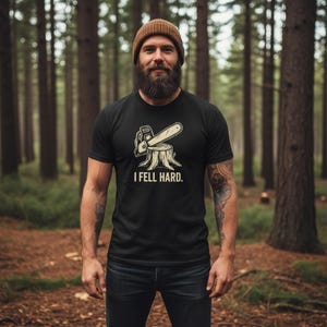 Pode incluir: Camiseta preta com um gráfico de motosserra em um tronco de árvore e o texto "I FELL HARD". A pessoa que usa a camiseta está em uma floresta. Ele está usando um gorro marrom e jeans.