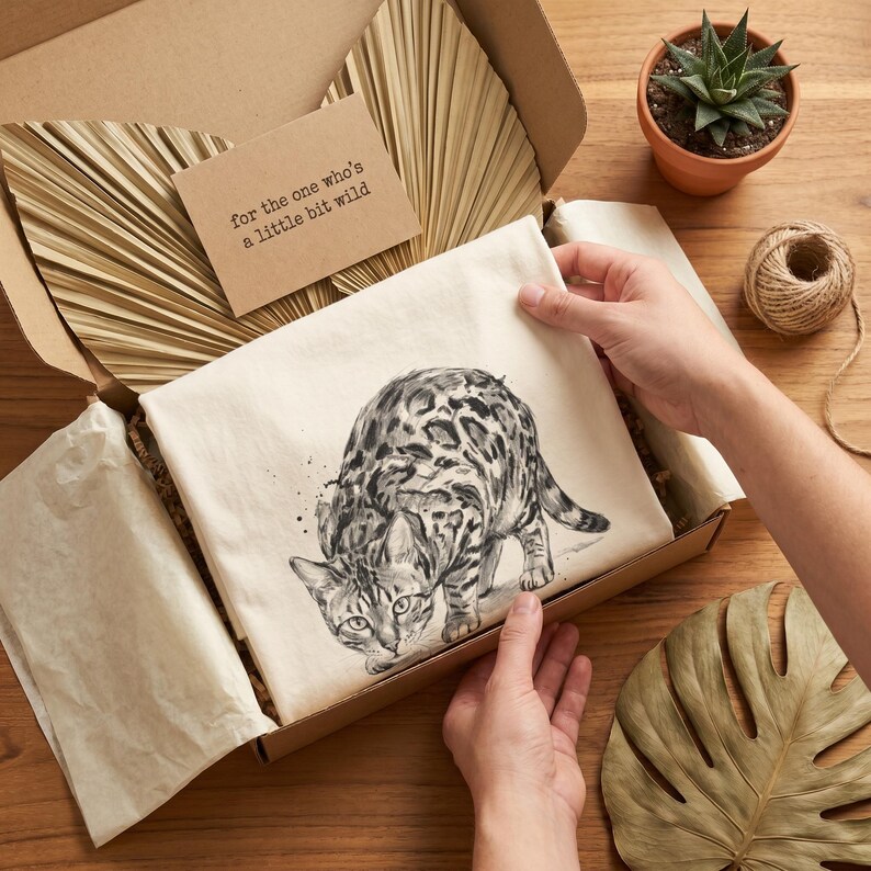 May include: A Bengal cat illustration on a cream-colored fabric, presented in a cardboard box. The cat is depicted in black and white with a spotted pattern. A small card with the text "for the one who's a little bit wild" is included.