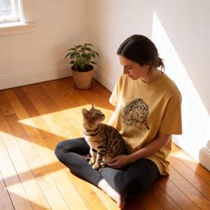 May include: Mustard yellow t-shirt with a graphic of two cats. A brown and black Bengal cat sits on a person's lap. The person is wearing black leggings and sitting on a wooden floor. A potted plant is in the background.
