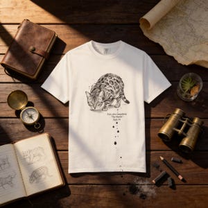 May include: White t-shirt featuring a detailed illustration of a Bengal cat in a stalking pose. The graphic is in black and white with the text "Felis catus bengalensis: The Hunter, Sept 19". The shirt is displayed on a wooden surface with a compass, journal, and binoculars.