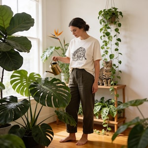 May include: A cream-colored t-shirt with a black illustration of a cat, paired with dark green pants. A brass watering can waters a large plant with green leaves. A tabby cat sits on a wooden shelf surrounded by plants. The room has a bright, natural light.