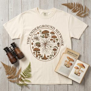 Op de afbeelding: Cr&egrave;mekleurig T-shirt met een cirkelvormige afbeelding van verschillende paddenstoelen en de tekst "THE UNDERGROUND NETWORK". Het shirt wordt getoond met een vintage boek getiteld "Mushrooms of North America" en een antieke verrekijker.