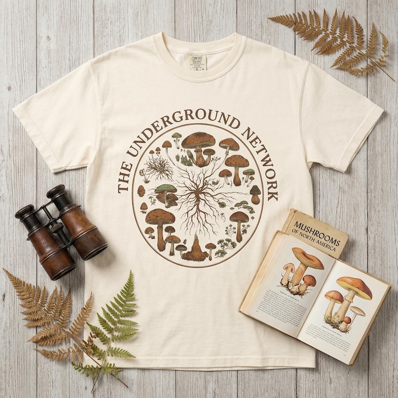 Op de afbeelding: Cr&egrave;mekleurig T-shirt met een cirkelvormige afbeelding van verschillende paddenstoelen en de tekst "THE UNDERGROUND NETWORK". Het shirt wordt getoond met een vintage boek getiteld "Mushrooms of North America" en een antieke verrekijker.