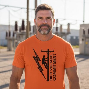 May include: Orange t-shirt with a black graphic of a lightning bolt, a utility pole with a lineman, and the text "HIGH VOLTAGE COMMITMENT." The shirt is short-sleeved and worn by a person.