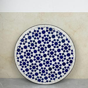 Może przedstawiać: Okrągły stół dekoracyjny z blatem z mozaiki. Płytki są białe i niebieskie, ułożone we wzór gwiazd. Stół ma czarną obwódkę i stoi na neutralnym tle. Średnica ok. 60 cm.