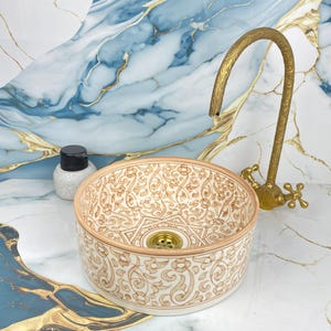 May include: A decorative ceramic sink with a beige and brown floral pattern. The sink is round with a gold drain and is paired with a vintage-style brass faucet. A small bottle with a black cap sits nearby.