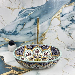 Puede incluir: Un lavabo ovalado de cerámica con un detallado estampado floral en azul, amarillo y rojo. El lavabo tiene un borde verde claro y un grifo dorado. A la izquierda del lavabo hay una pequeña botella blanca con tapa negra.