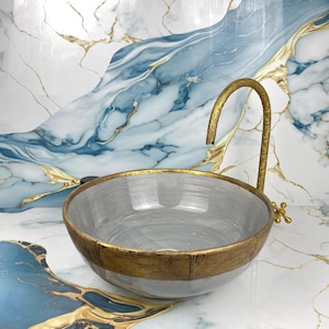 Peut inclure: Un lavabo en forme de vasque gris et doré avec un robinet doré incurvé. Le lavabo a une forme de bol rond avec une bande dorée autour du bord. Le fond présente un motif de marbre avec des accents bleus et dorés.