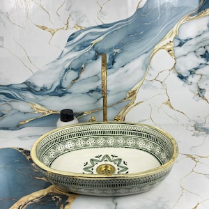 Peut inclure: Évier ovale en céramique avec un motif géométrique vert et blanc. L'évier a un bord doré et un robinet assorti. Une petite bouteille avec un bouchon noir est à proximité. Le fond présente un motif de type marbre avec des accents bleus et dorés.