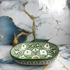 May include: An oval, green and white ceramic sink with a floral pattern. The sink has a gold tap and is set against a marble-patterned wall. A decorative gold metal vase is to the left of the sink.
