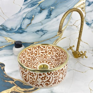 Peut inclure: Un lavabo décoratif en céramique avec un motif floral marron et blanc et un bord doré. Il est associé à un robinet en laiton de style vintage. Une petite bouteille blanche avec un bouchon noir est également visible.