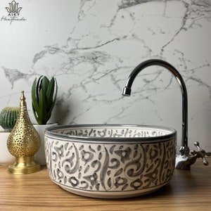 May include: A white ceramic sink with a gray floral pattern. The sink is round and has a wide rim. A chrome faucet is mounted on the wall behind the sink.