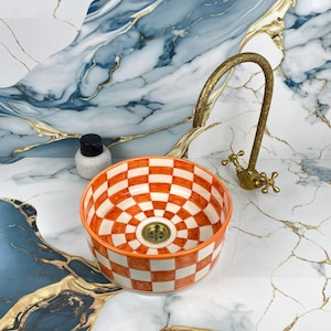 Puede incluir: Un lavabo redondo de cerámica a cuadros naranja y blanco con un grifo de latón. El lavabo tiene un desagüe central y está sobre un fondo de mármol. También se ve una pequeña botella blanca con tapa negra.