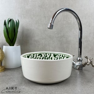 Pode incluir: Pia de cerâmica branca com um design estampado em verde e branco. A pia é redonda e tem uma borda larga. Uma torneira cromada é visível no fundo.